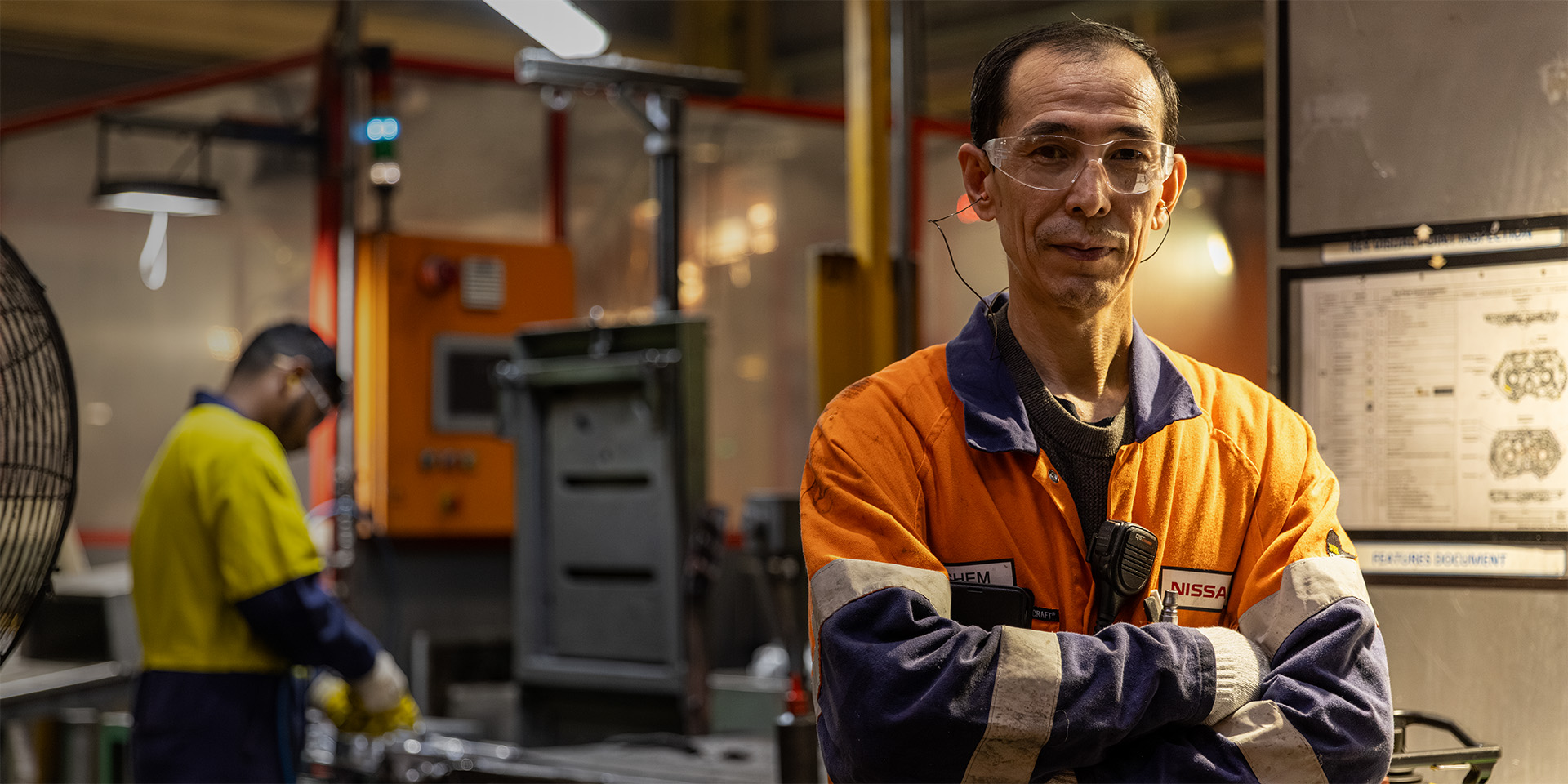 A worker in hi-vis overalls and safety glasses standing with arms folded on a factory floor, a colleague at a machine behind
