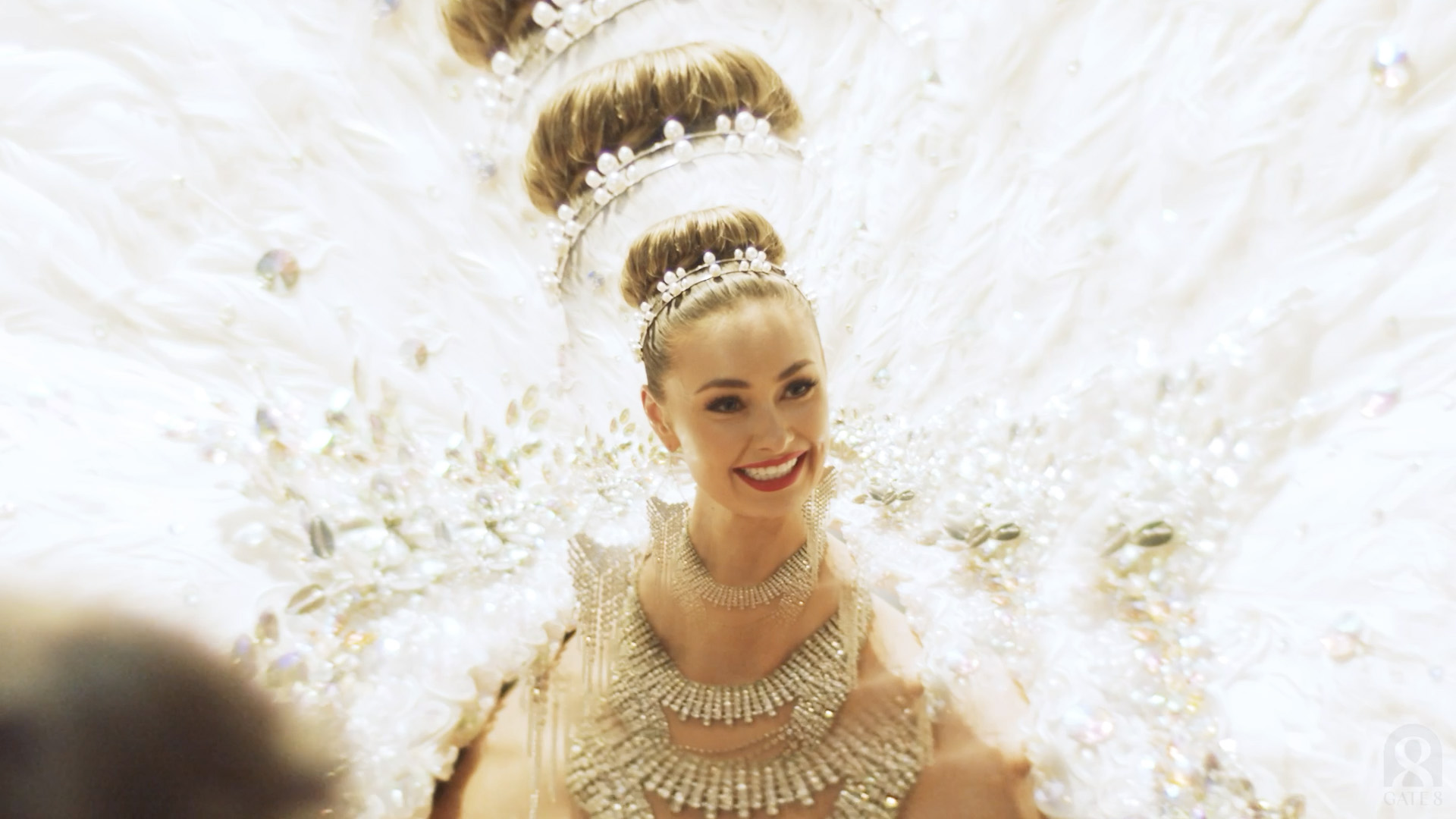A performer in a white feathered costume and crystal headdress, smiling to camera