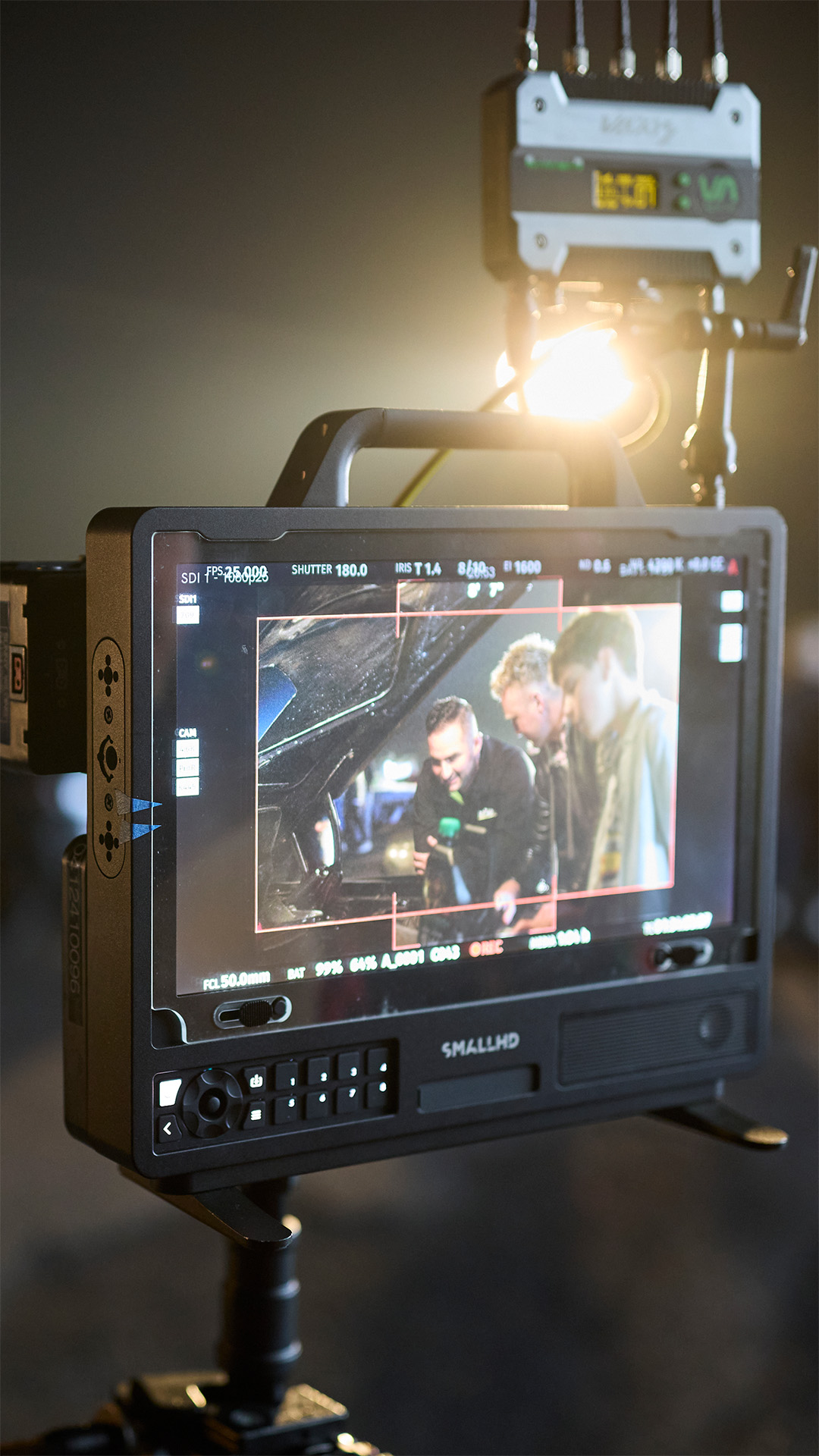 An on-set monitor showing three people at an open car bonnet, framing guides overlaid, with a light and rigging above it