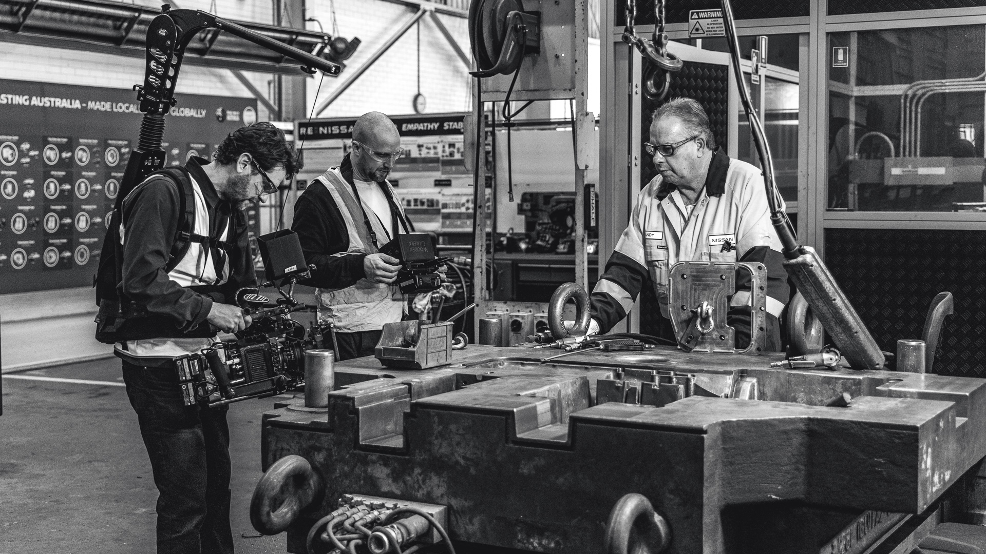 Black and white: a camera operator and a crew member in hi-vis filming a worker at a large industrial press on a factory floor